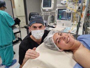 A smiling young woman lies in a hospital bed after surgery, wearing a hairnet and hospital gown, while a masked surgeon kneels beside her