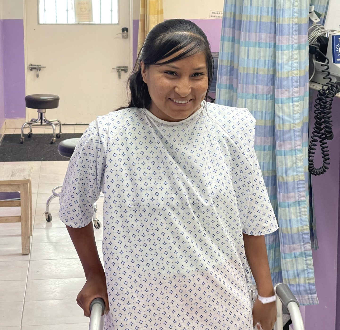 The same Tarahumara woman smiling and using a walker in a hospital room after surgery, wearing a hospital gown, showing signs of healing and hope.